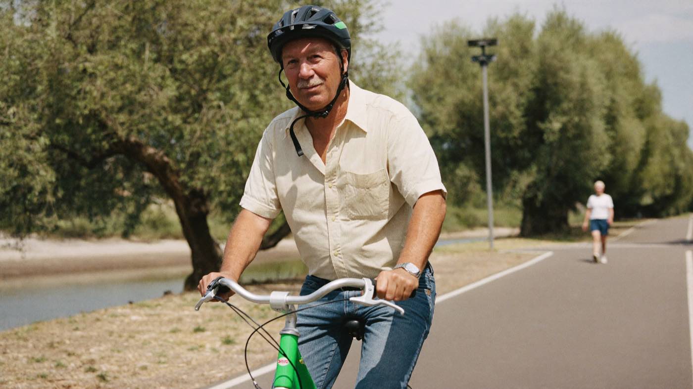 A man in his late sixties riding a bicycle at an easy pace along a flat path beside a river, with a walker further along the path behind him.