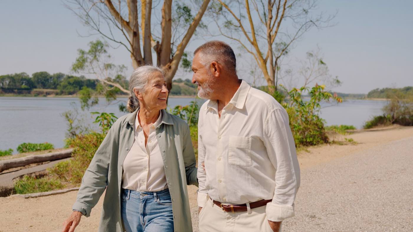A woman and a man in their late sixties walking side by side along a river path in the morning sun, mid-conversation.