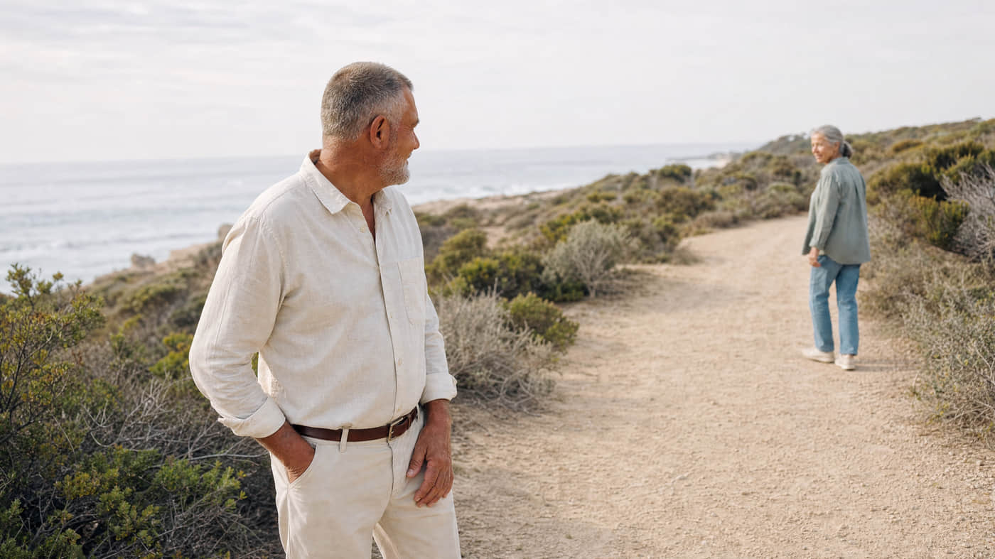 A man in his late sixties has stopped part-way along a coastal path, one hand resting at his hip and his weight shifted off that leg, while a woman of similar age walks on ahead and half-turns back to wait for him.