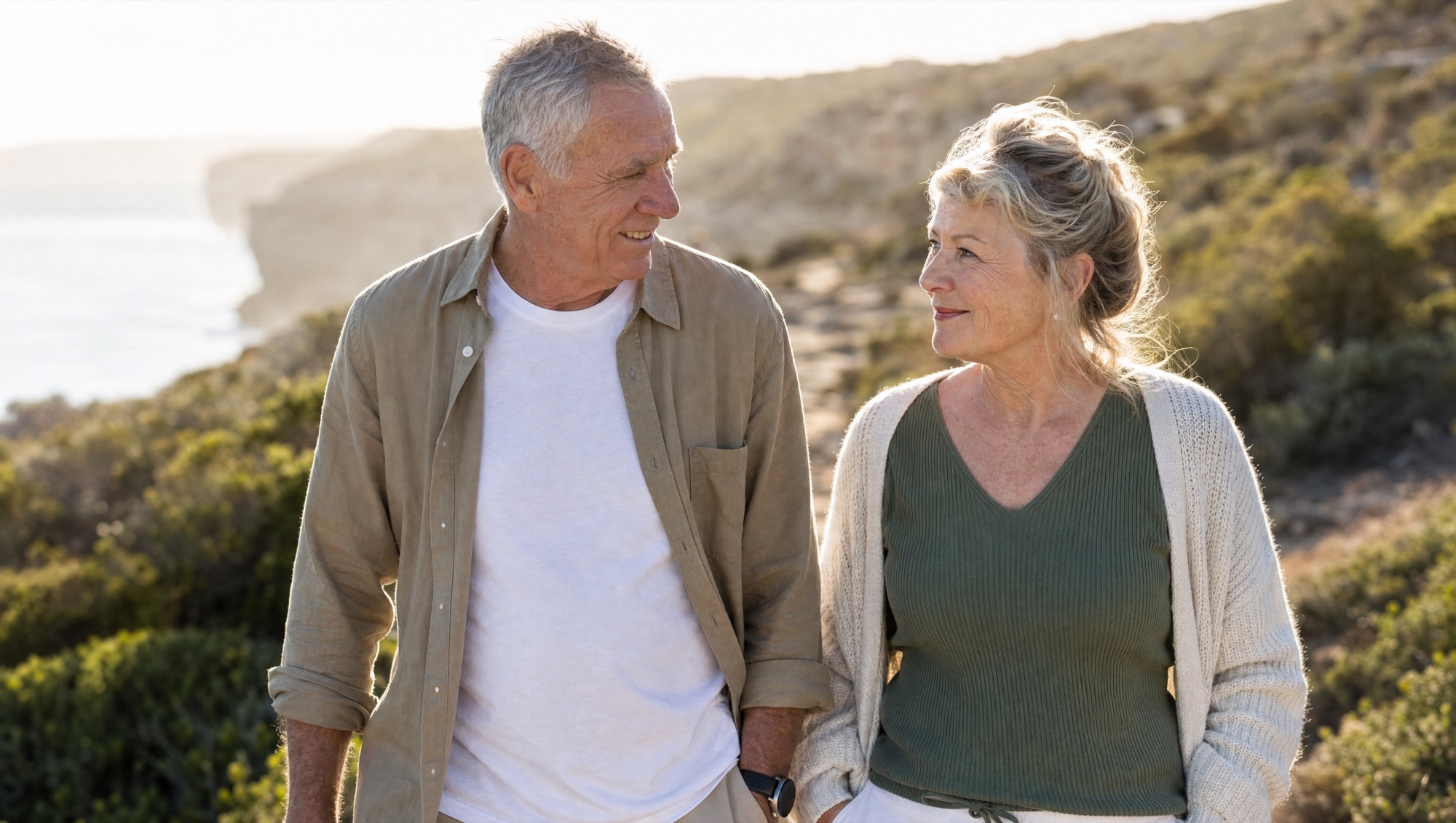 A man and a woman in their late sixties walking together along a coastal path in the morning sun.