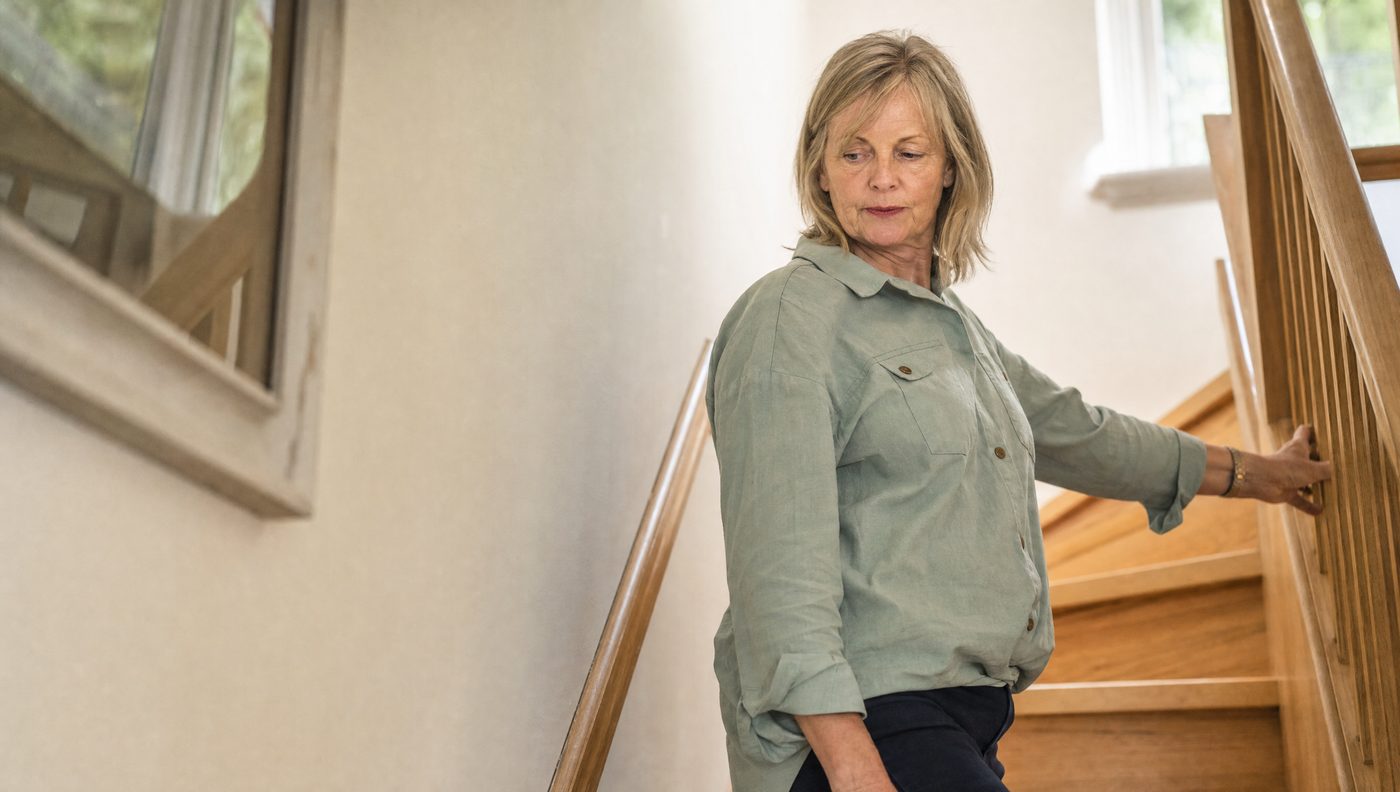 A woman in her sixties pausing on a staircase with one hand on the handrail.