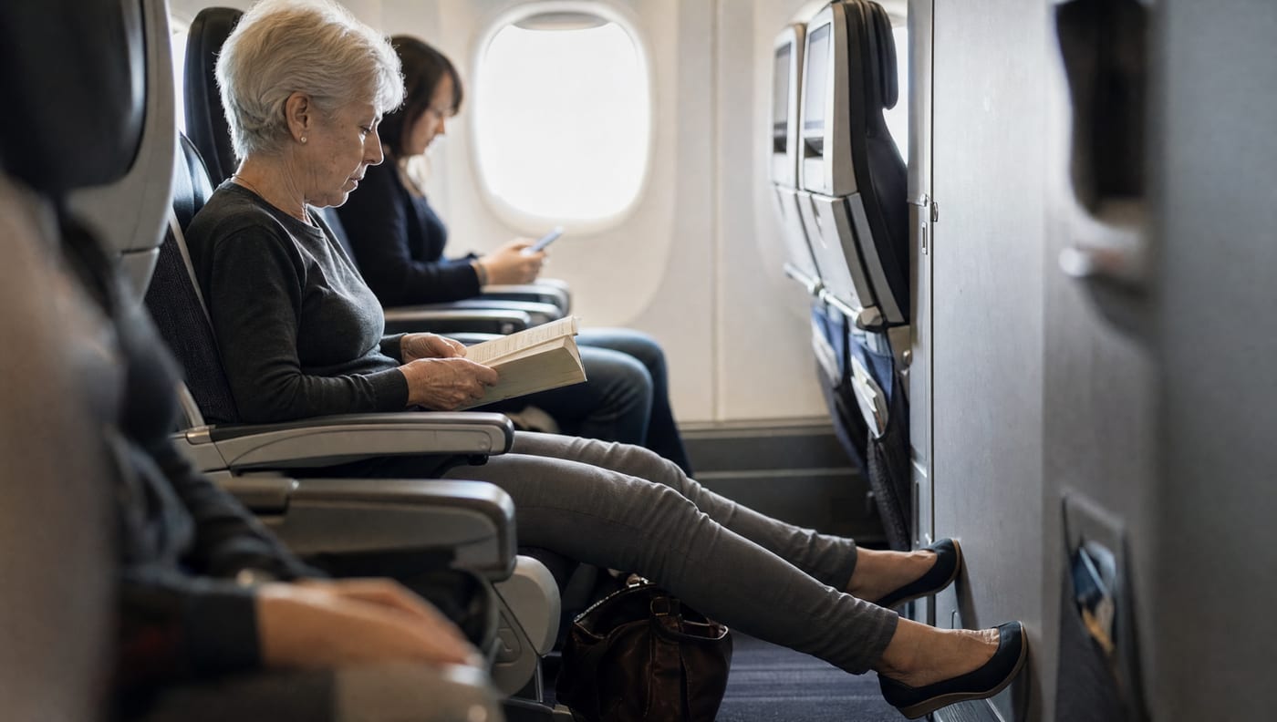 A woman in her late sixties seated in the aisle seat of an aircraft, one leg stretched into the clear aisle with the foot flat on the floor, reading a paperback.