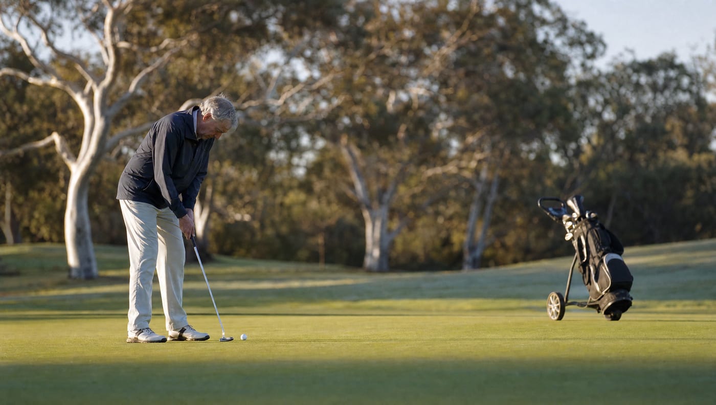 A man in his sixties putting on a golf course green in early morning light, a pushcart with a golf bag parked behind him.